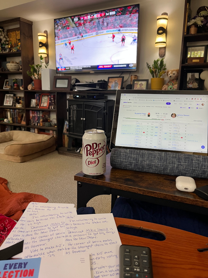 A view from my sofa. Background: a NJ Devils hockey game on the TV; middle ground: a tablet open to my fantasy football team and a Diet Dr. Pepper; foreground: postcards being written to NJ voters for Mikie Sherrill
