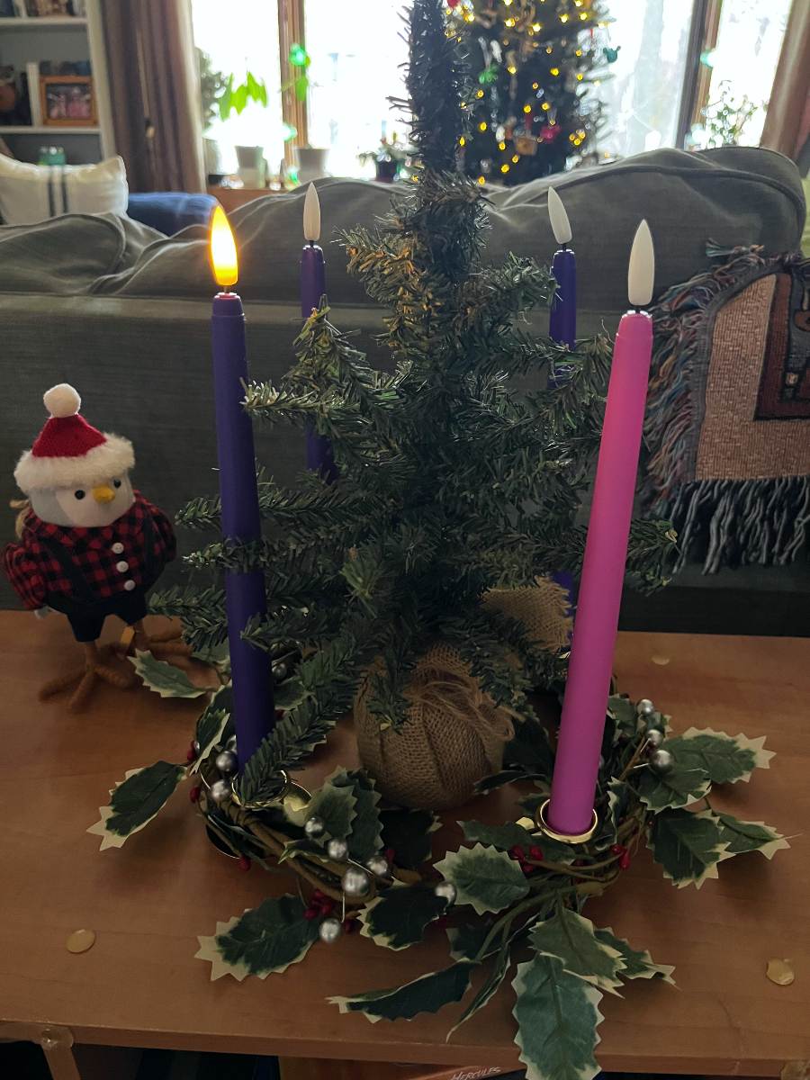a leafy advent wreath with 1 lit purple candle and the other 3 unlit. There is a small fake tree in the middle, and one bird figurine looking on.