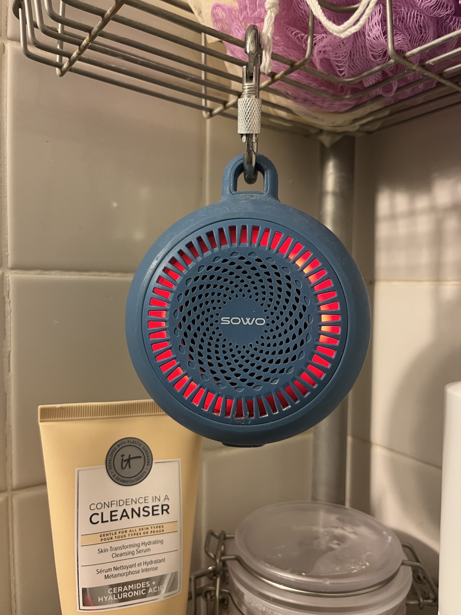 a photo of a sowo shower speaker hanging off of a shower shelf. My face cleanser - IT cosmetics Confidence in a Cleanser is also visible
