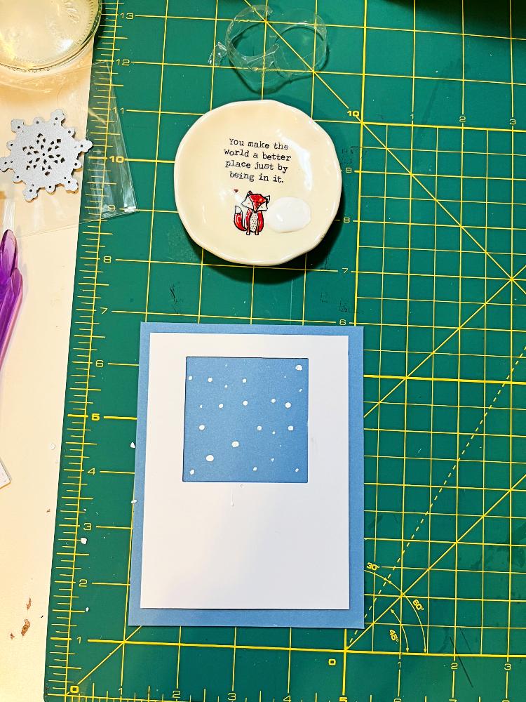 a top-down view of a round dish with white paint in it, and a card front with white dots showing through a square cutout