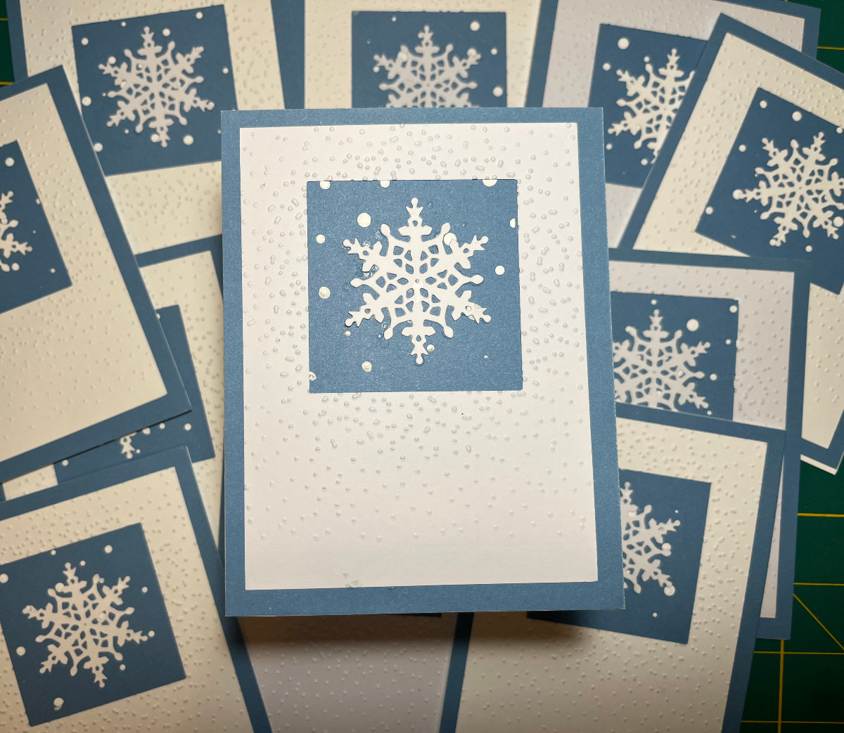 a card with a blue background, white panel with a square cutout. In that cutout are dotted paint snowflakes and one large paper snowflake.