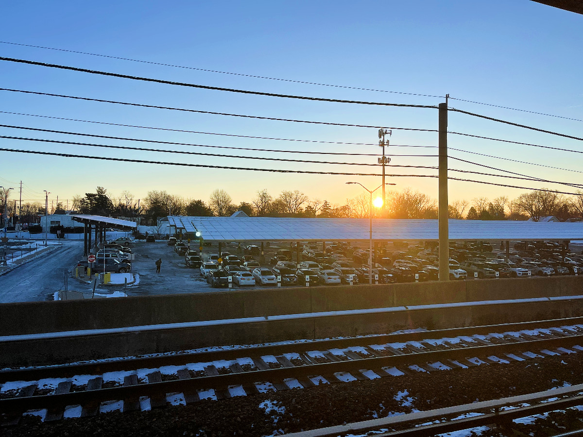 the sun is rising over a very chilly train station parking lot. there is snow on the solar panels that cover the lot. the entire photo looks cold.