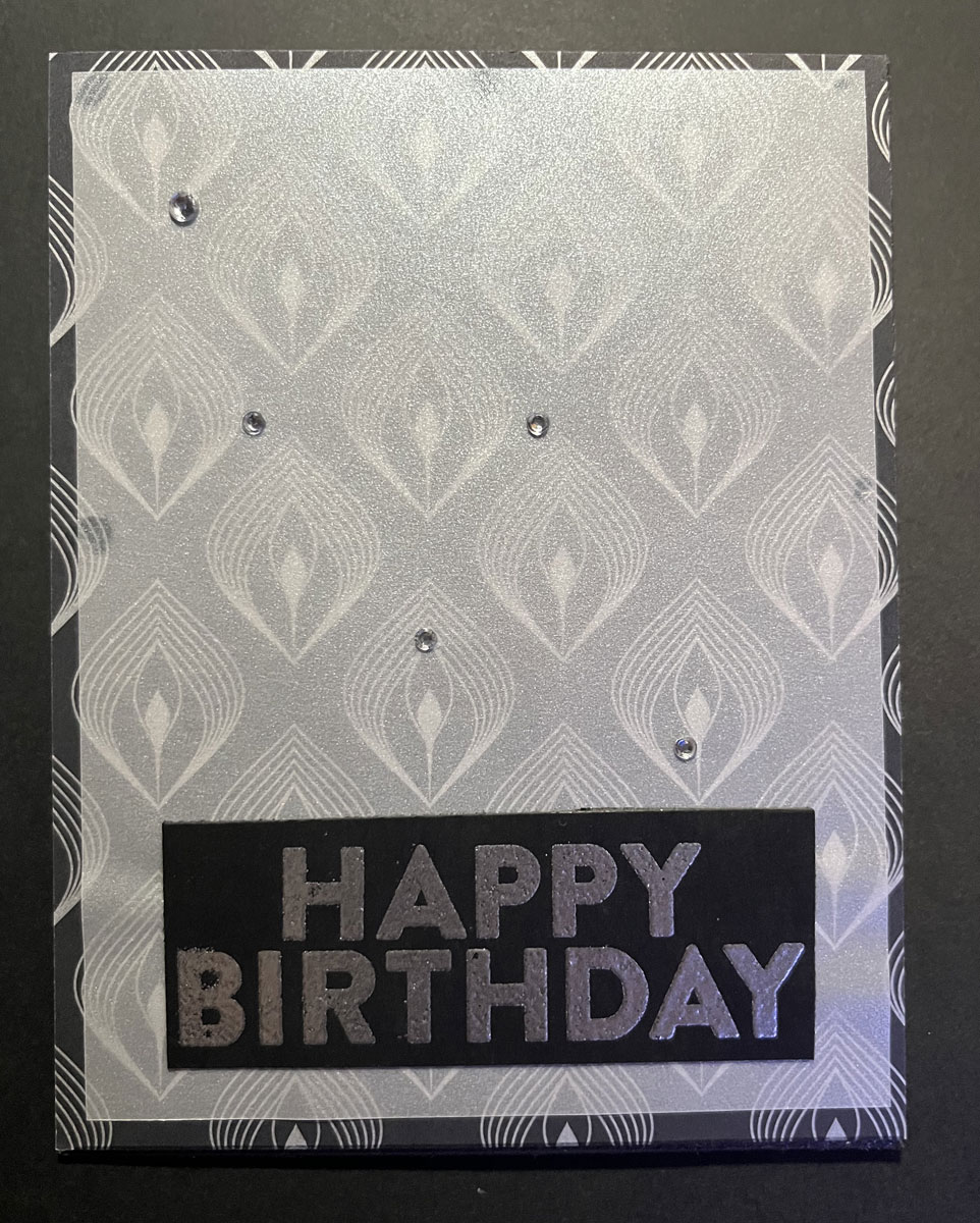 a card front, vellum on a black/silver diamond patterened background, with HAPPY BIRTHDAY embossed in silver onto black, and 5 tiny gems glued to it. it's art deco-ish to me.