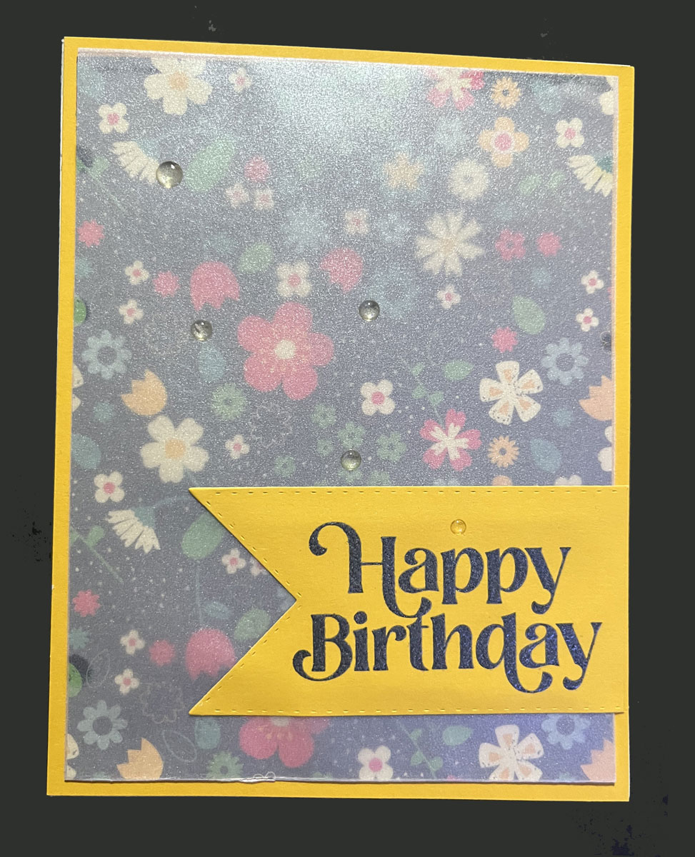 a birthday card; yellow background; various flowers on a black background, vellum over the printed paper, happy birthday embossed in black on a yellow piece of paper cut into a ribbon shape. 