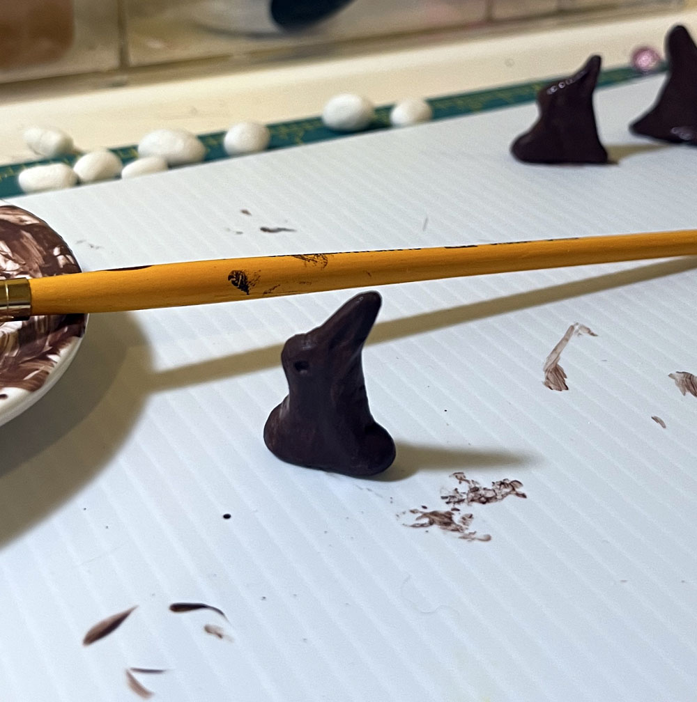 a brown painted bunny, with a dish of brown paint, a paintbrush, and some more chocolate rabbits in the background.