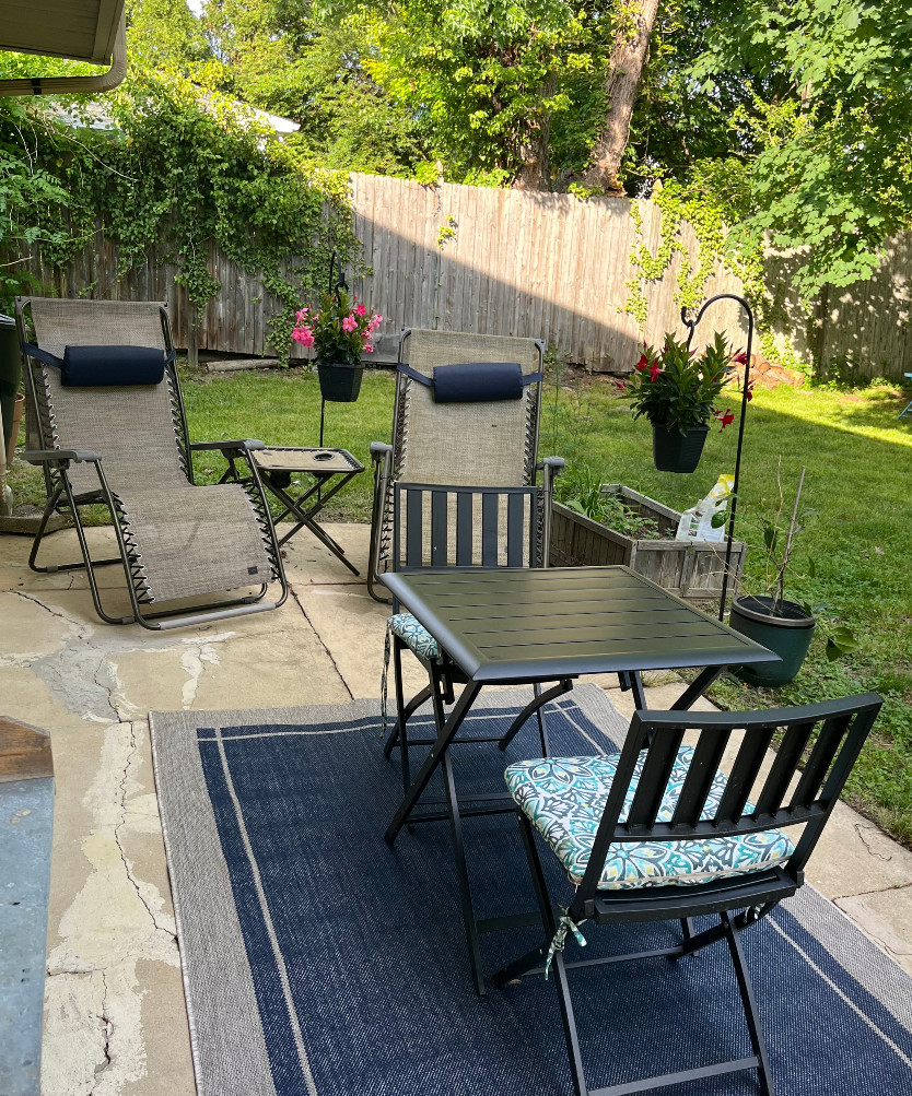my back slab set up for summer, some zero gravity chairs, a black bistro set on top of a blue outdoor carpet, pretty hanging flowers